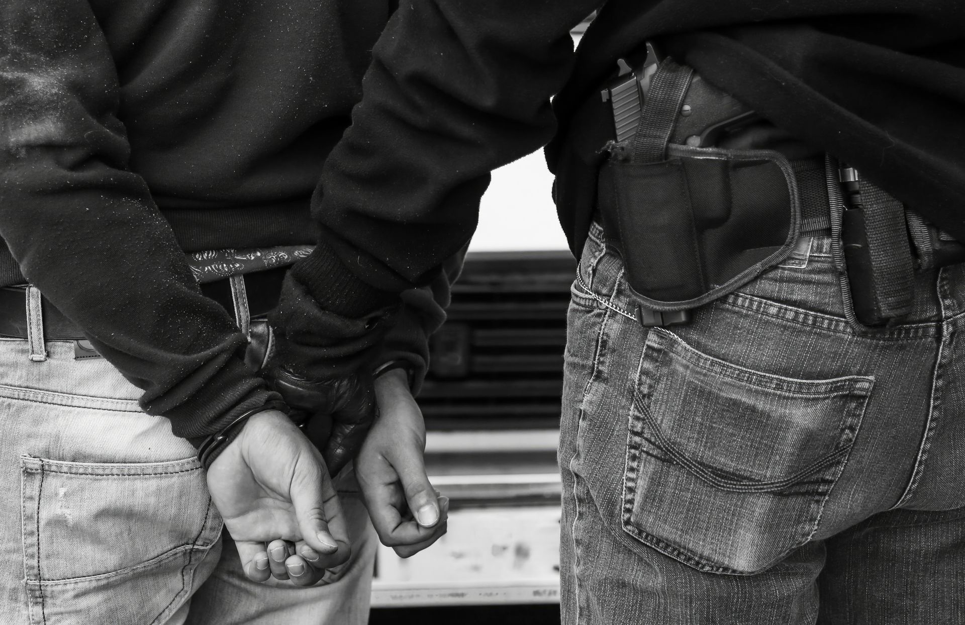 Person in handcuffs, being guarded by a person with a holstered gun.