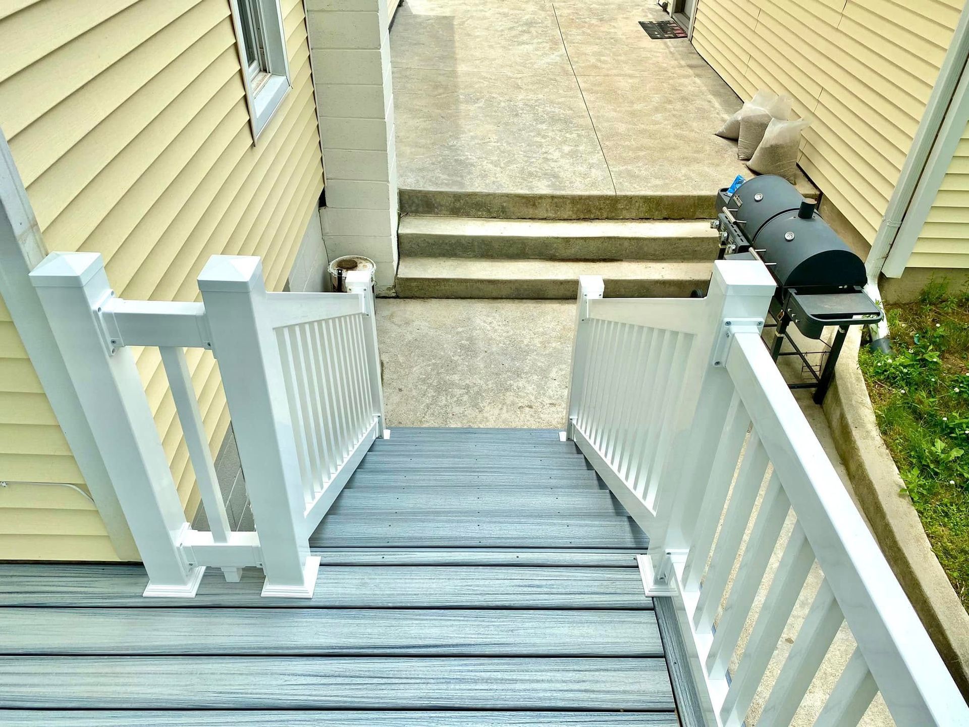 Wooden deck with white railing leads down to concrete stairs and a walkway. A grill is visible at the bottom.