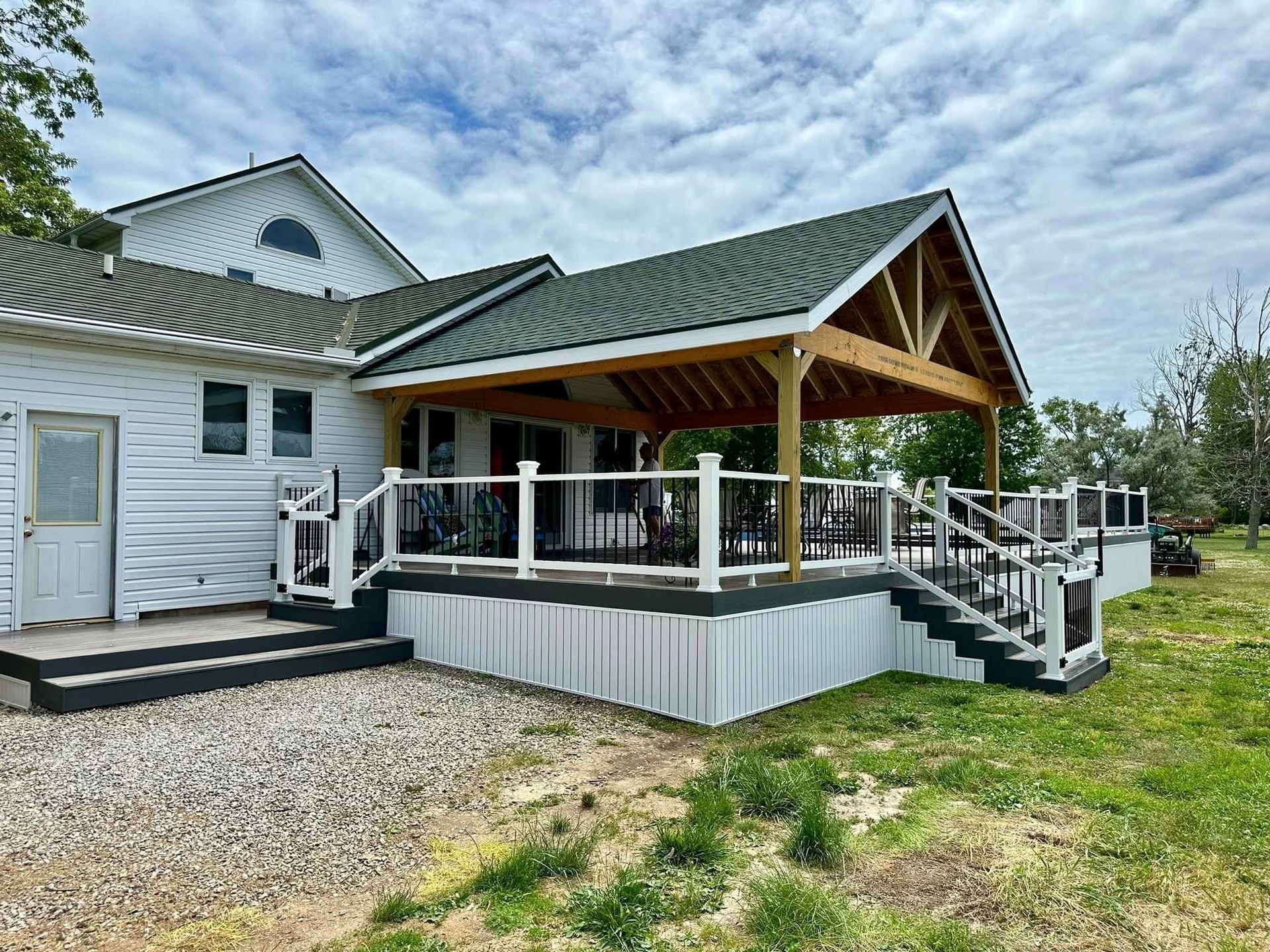 White house with a large deck and covered patio; green roof, wooden beams, railing, gravel, and grass.