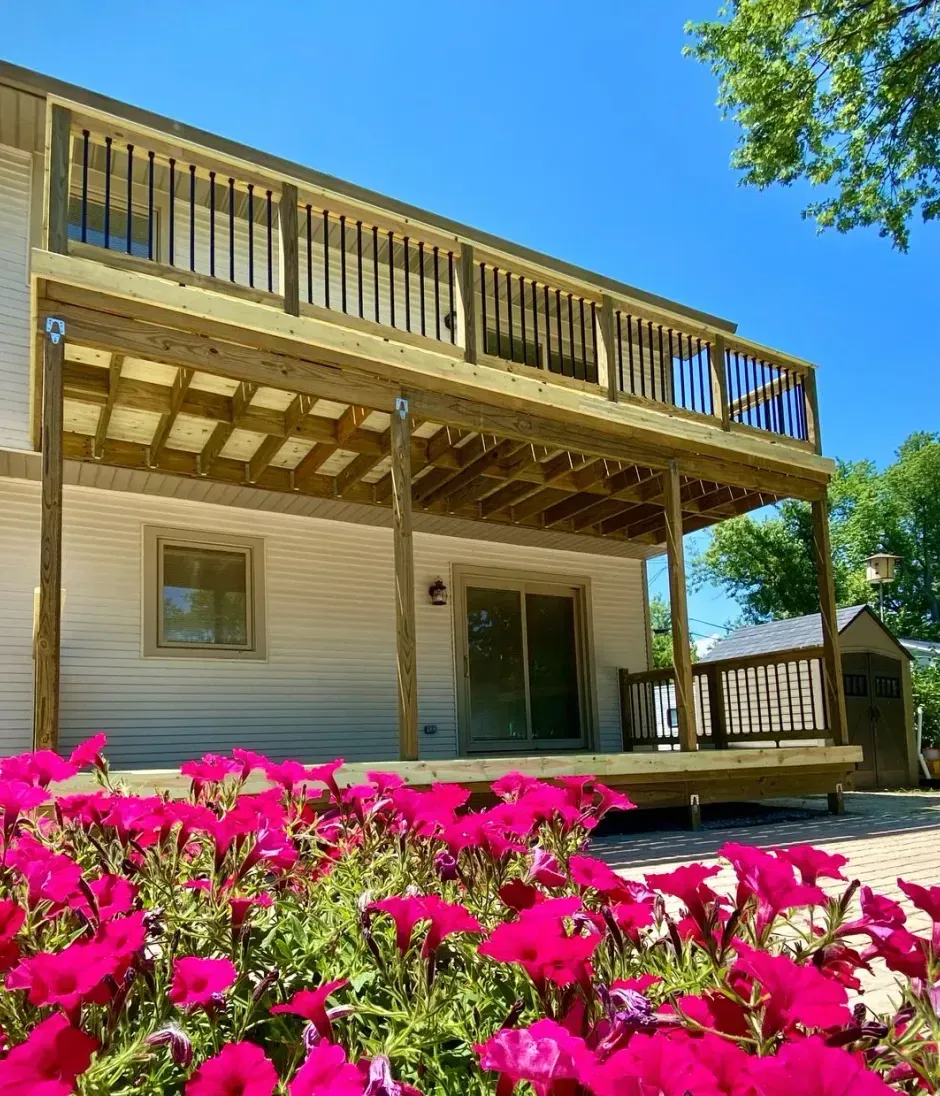 Two-story wooden deck attached to a light-colored house. Pink flowers in the foreground. Blue sky.