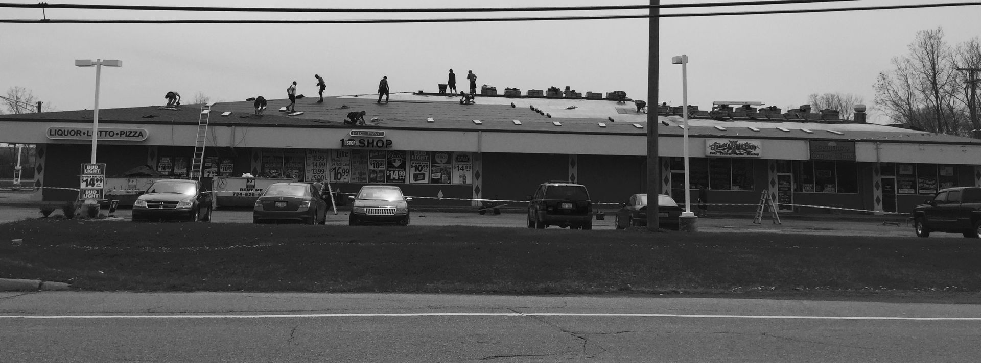 A strip mall with cars parked in front. People are on the roof. Gray sky.