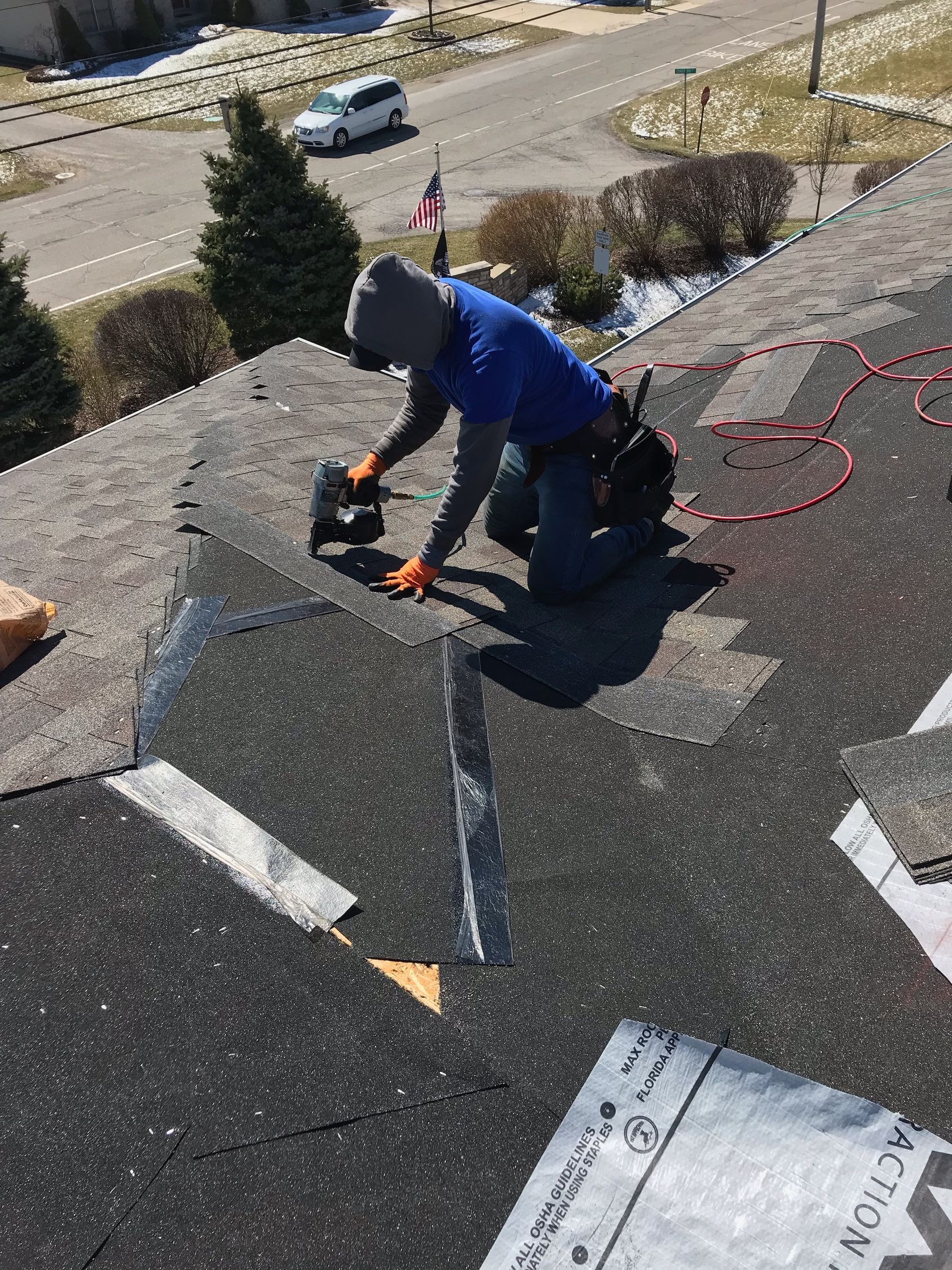 Roofer kneeling on a roof, using a nail gun. Blue shirt, orange gloves, and gray cap.