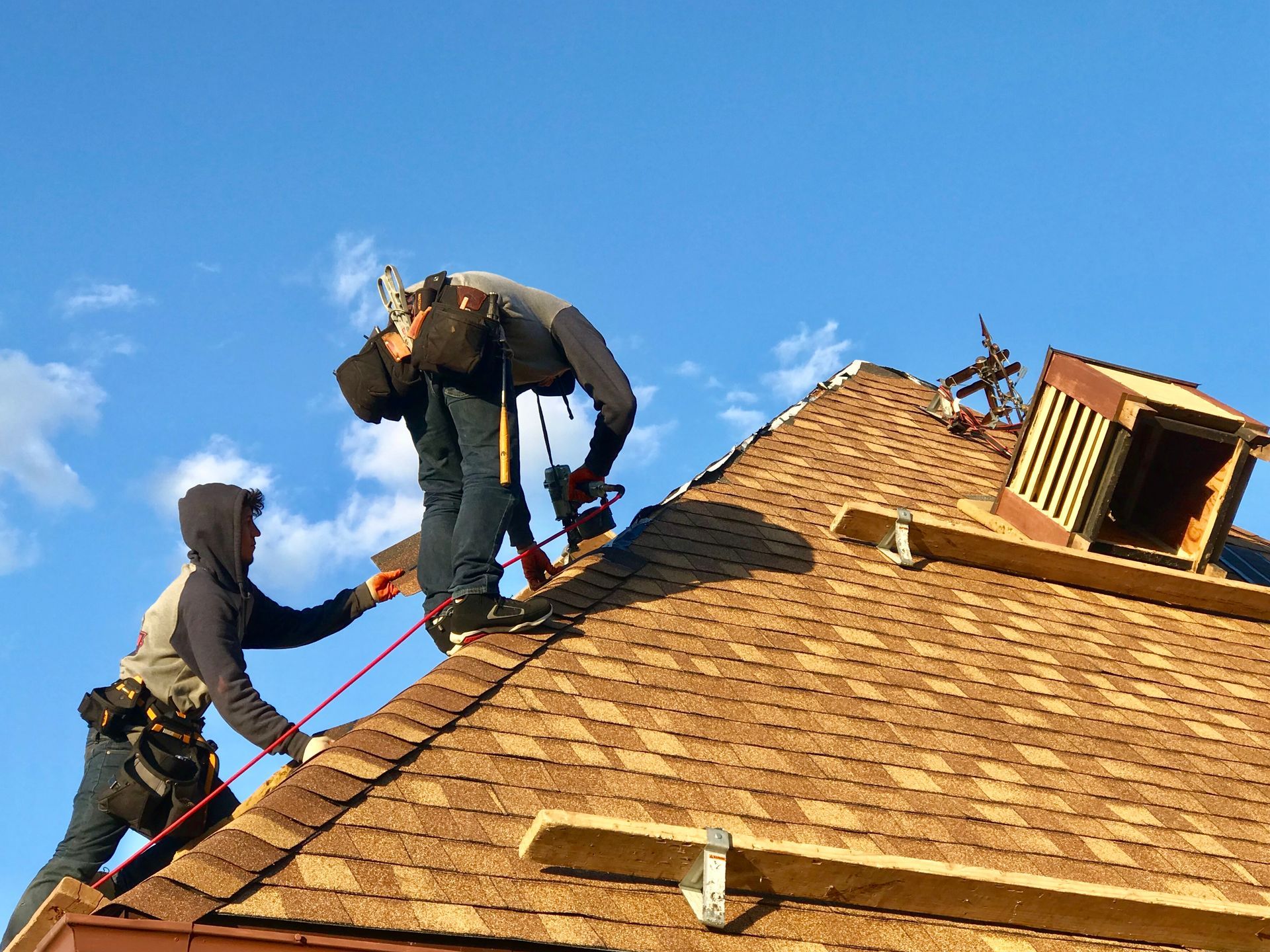 Two roofers working on a brown shingled roof under a blue sky. One is tethered to a safety line.