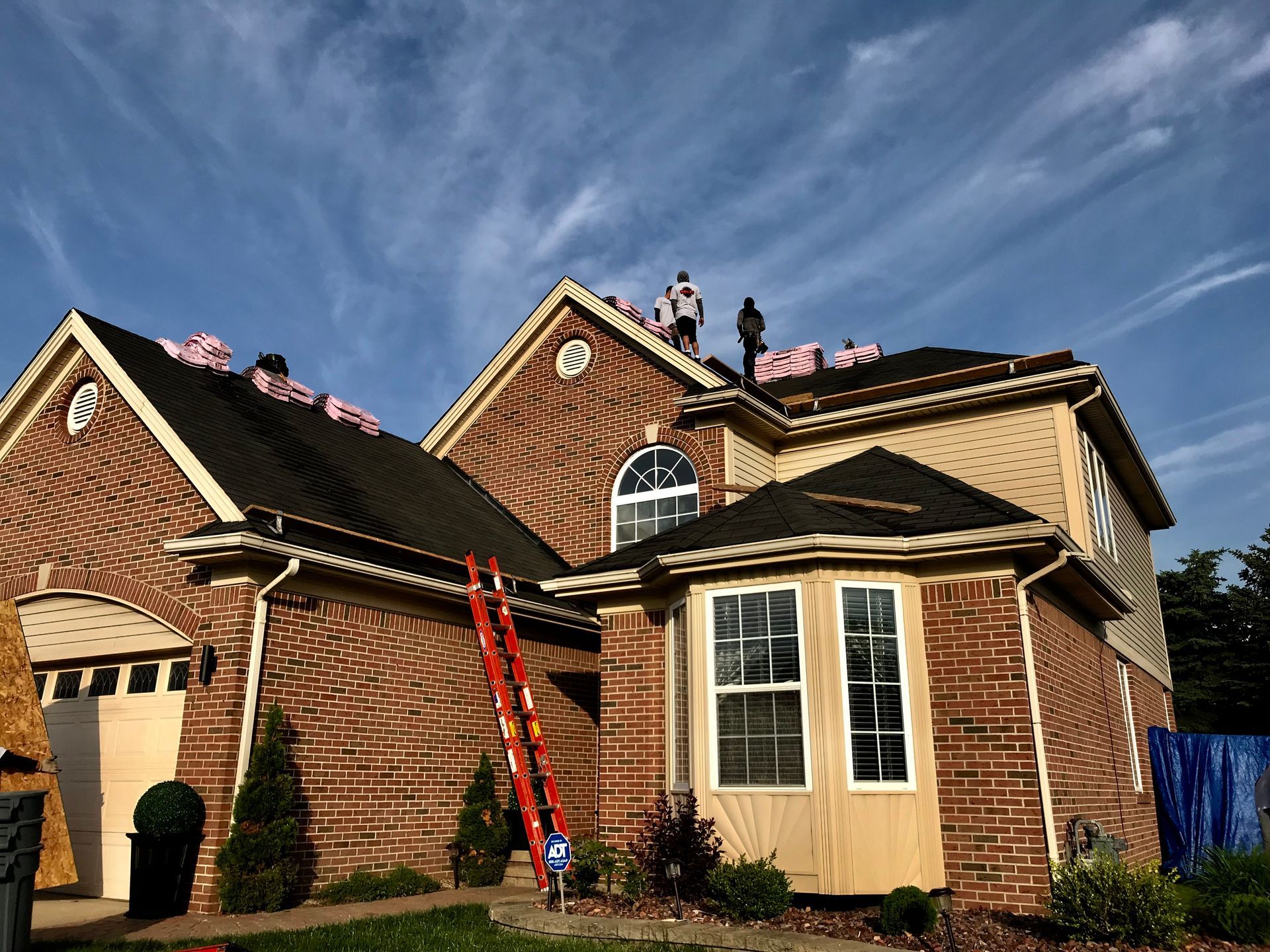 Roofers installing shingles on a two-story brick house with a red ladder and a blue tarp.