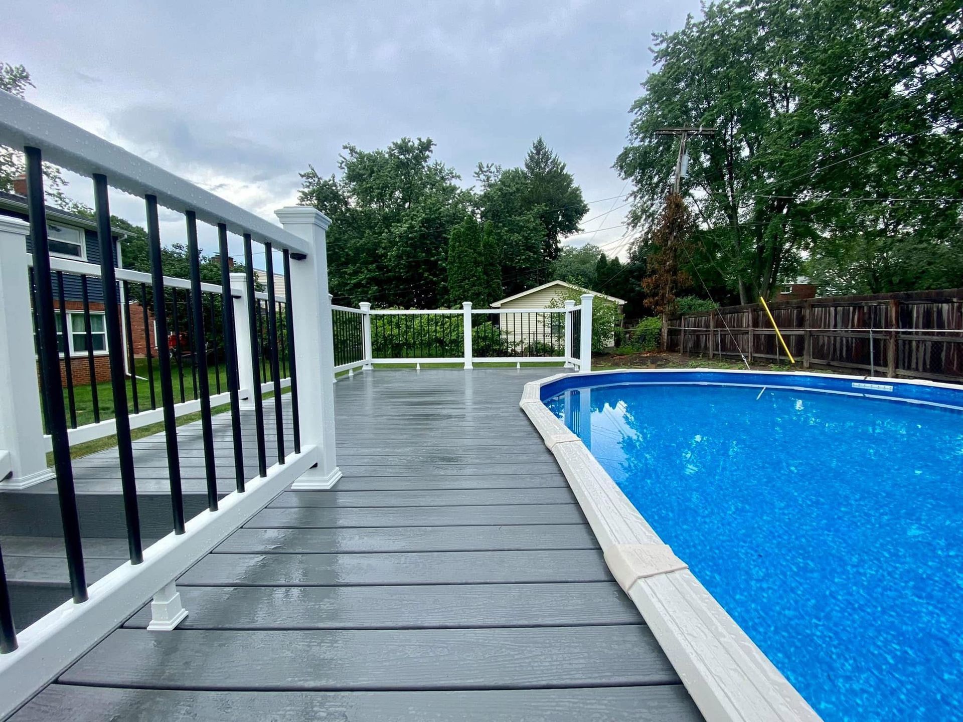 Gray deck alongside a blue swimming pool with white and black railing. Cloudy sky and trees in the background.
