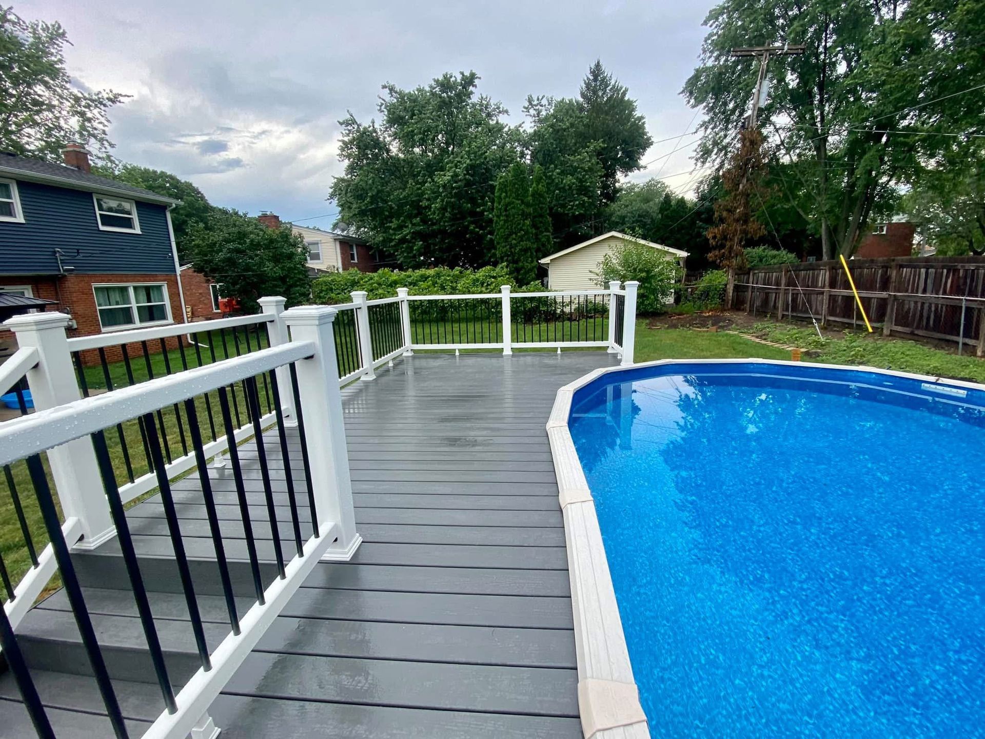 Gray deck with white railing next to blue swimming pool in a backyard.