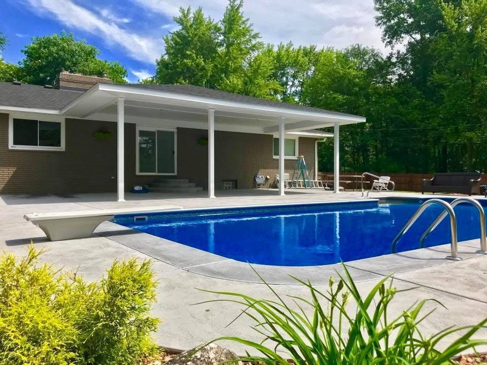 Poolside view: blue pool with diving board, concrete patio, white patio cover, brown house, lush green trees.