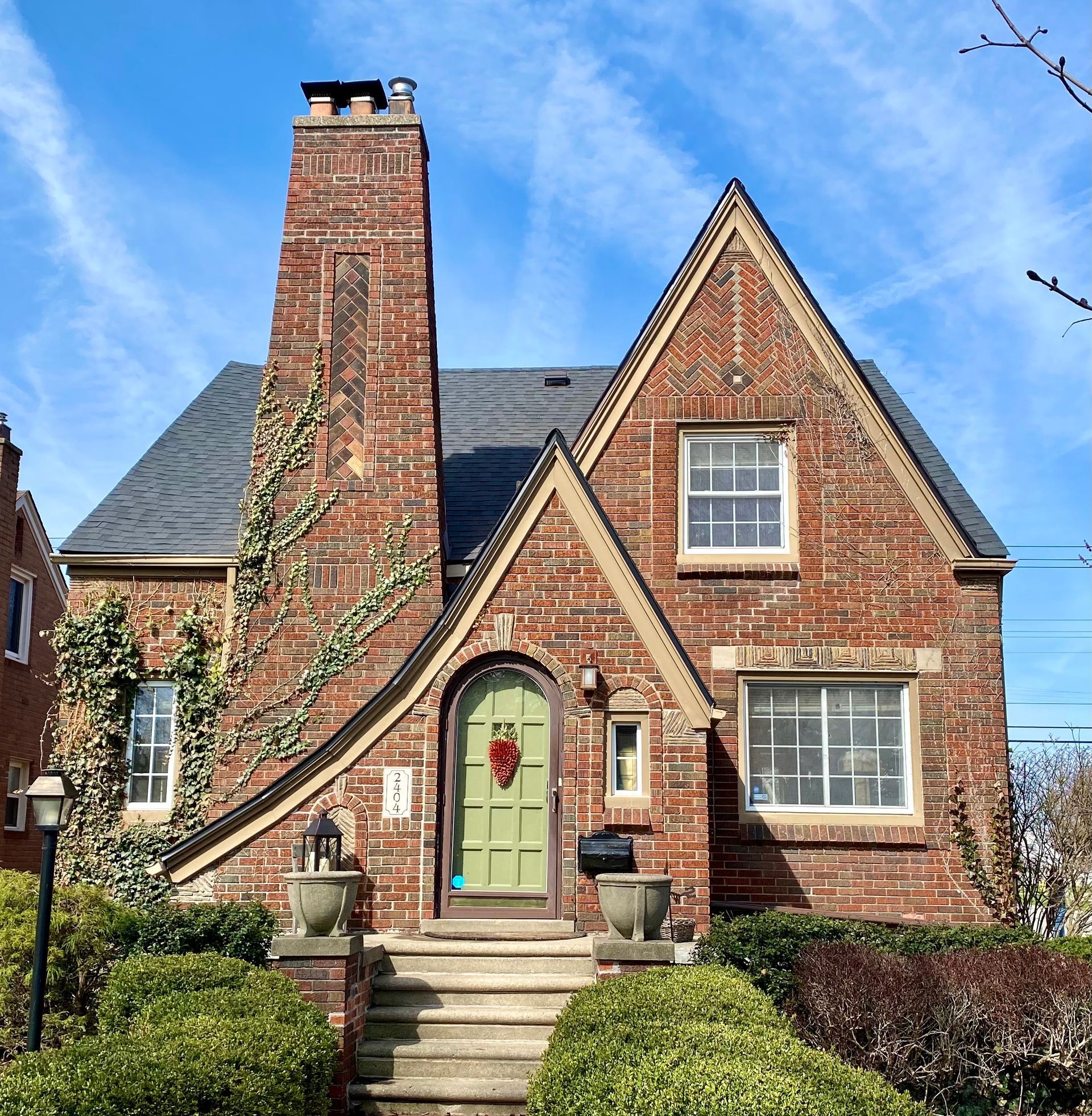 Brick Tudor-style house with green door, prominent chimney, and landscaping under a blue sky.