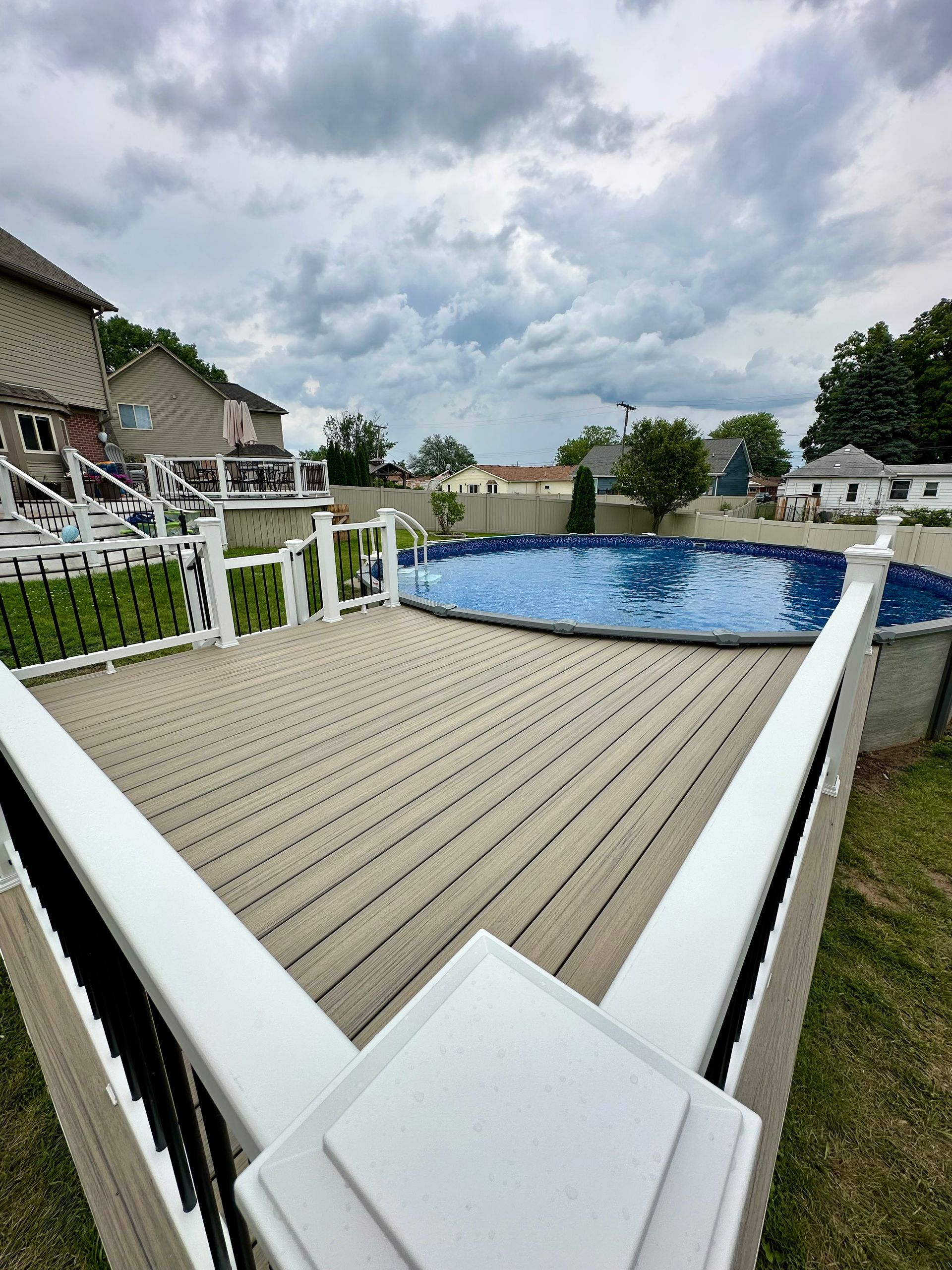 A deck surrounds a blue above-ground pool, with houses and a cloudy sky in the background.