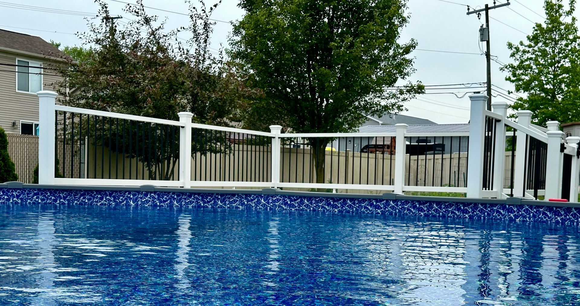 Pool with blue tiled surface, white and black fence, green trees, and cloudy sky.