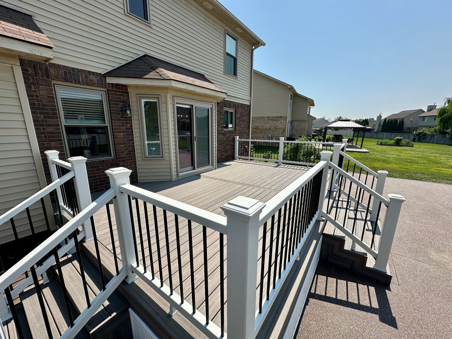 A two-level wooden deck with white railings and black spindles, attached to a two-story beige house with a brick accent.