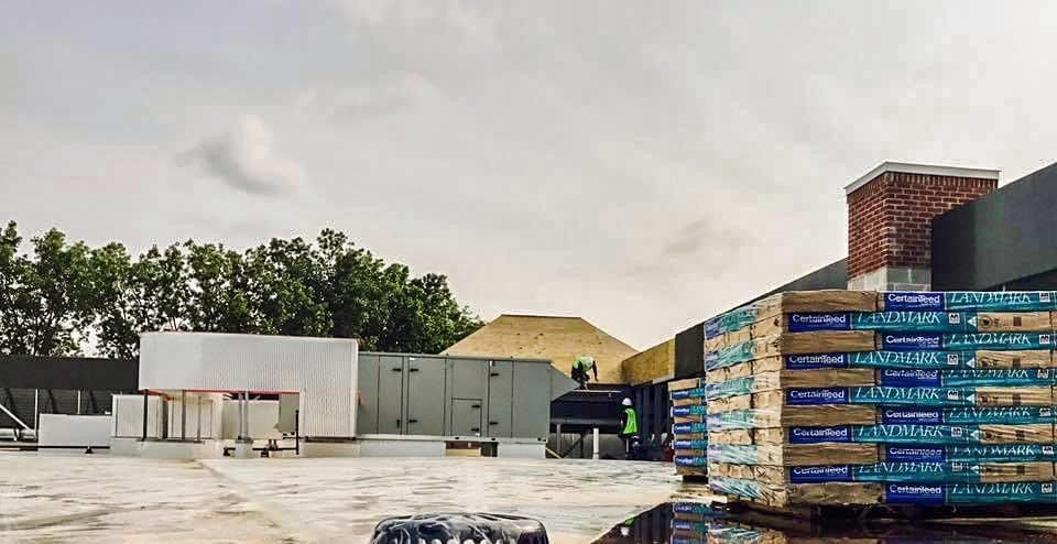 Pallets of materials stacked on a roof, with utility structures and trees in the background under a cloudy sky.