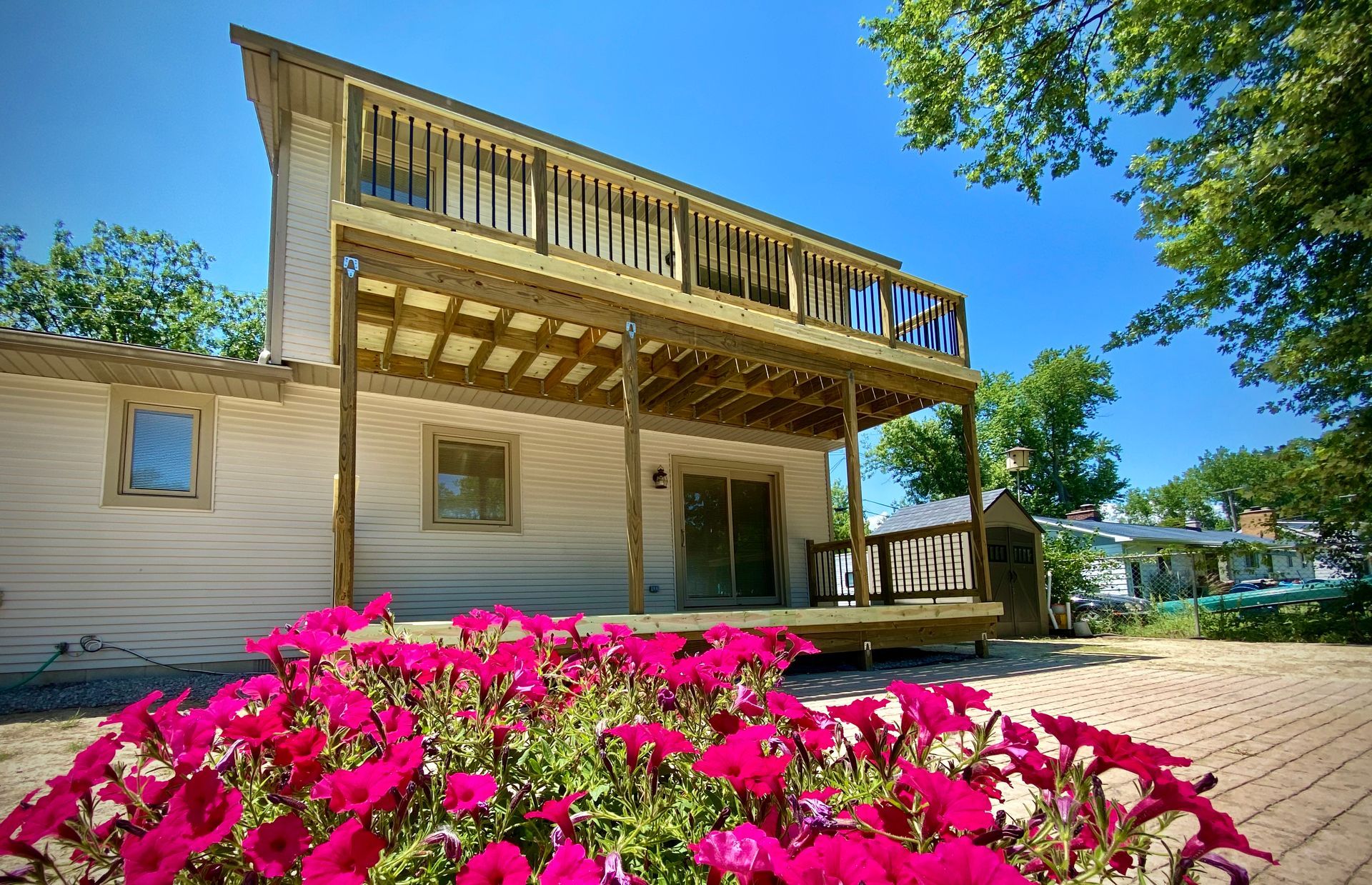 Two-story house with a wooden deck and balcony, red flowers in the foreground, sunny day.