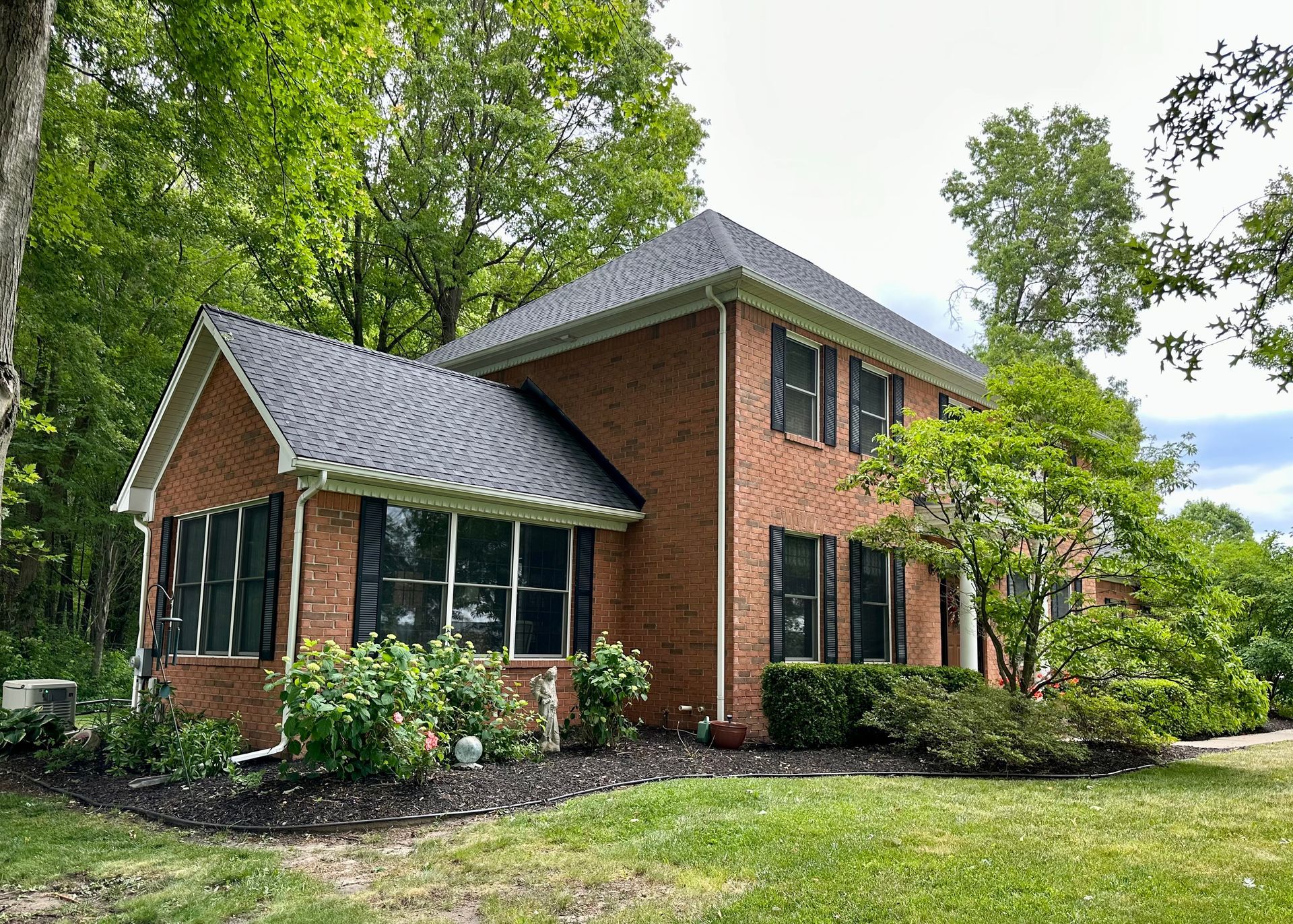 Two-story brick house with black shutters and roof, surrounded by trees and landscaping.