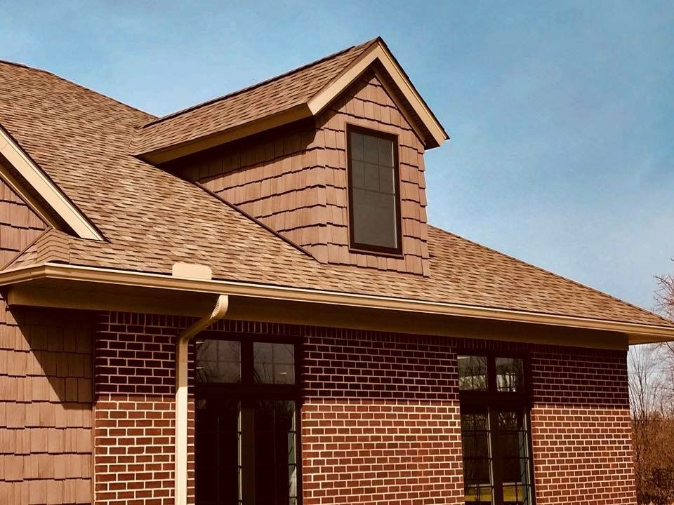 Brick house with brown roof and shingle siding, dormer window, and black framed windows.