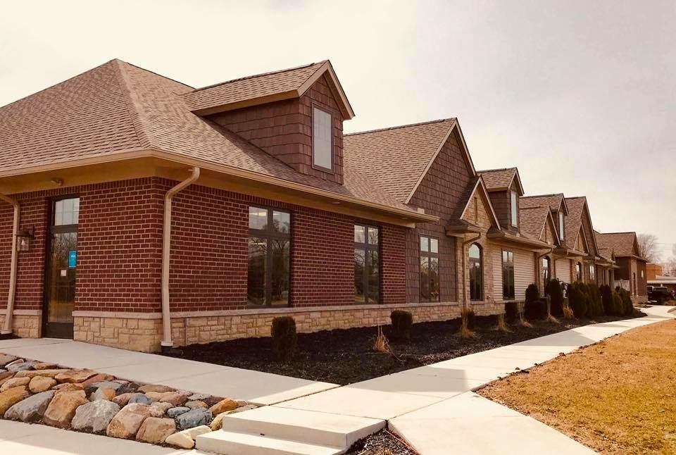 Row of brick and stone commercial buildings with multiple gabled roofs, a sidewalk, and landscaping.