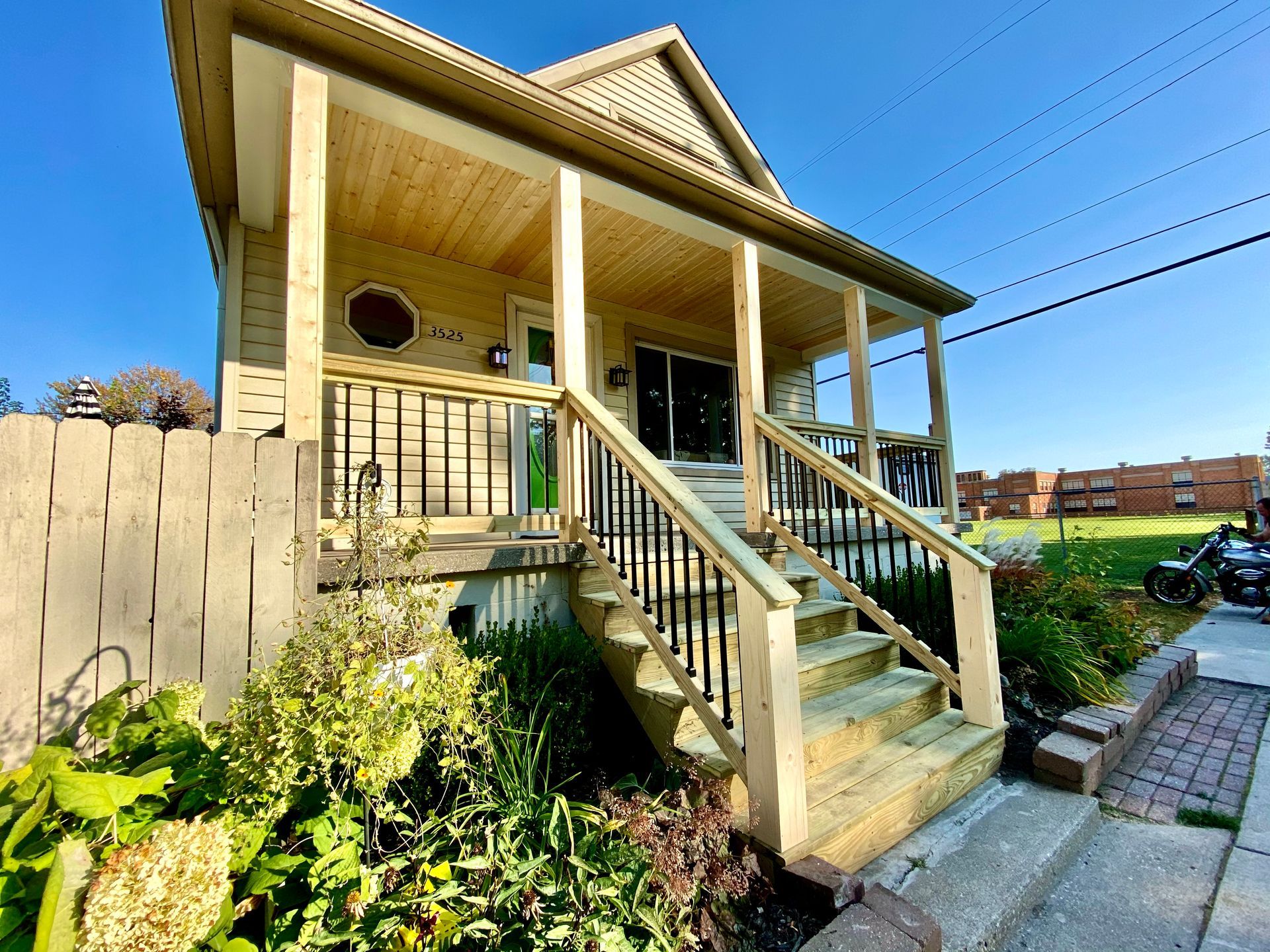 House with wooden porch, steps, and black railing. Green door, blue sky, and foliage.