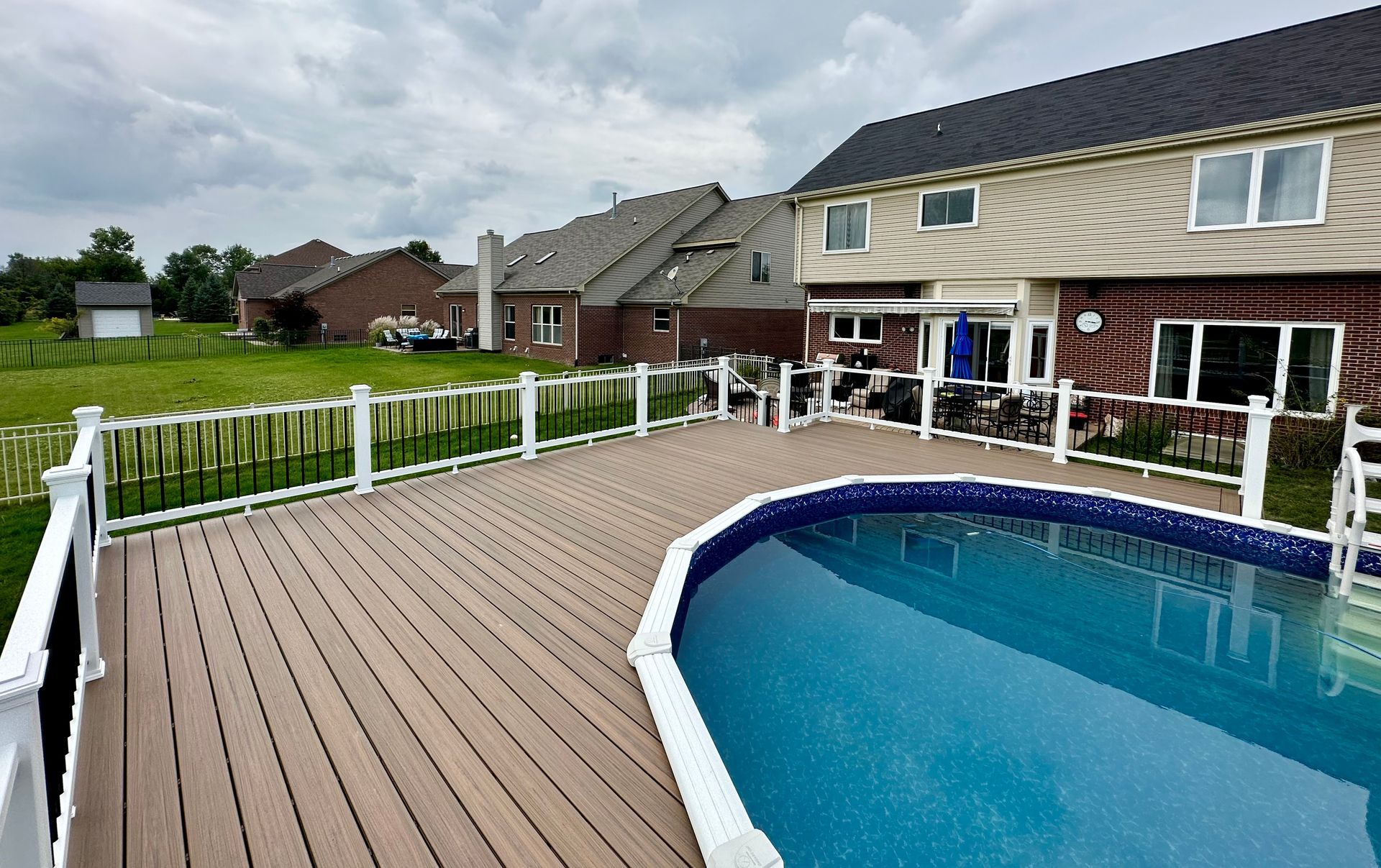 Backyard with a pool, deck, fence, and a two-story house under a cloudy sky.