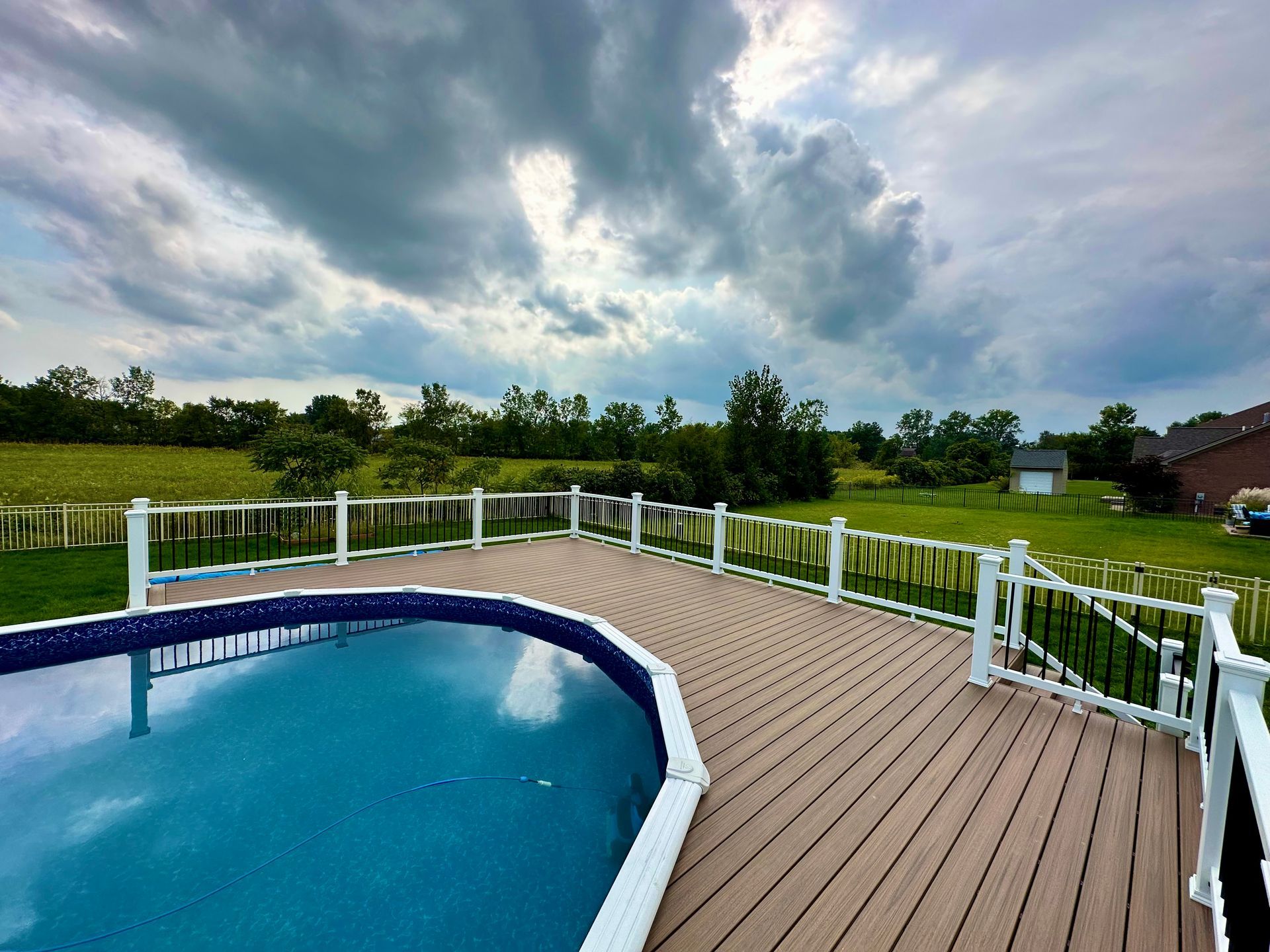 Pool deck with brown decking, white railing, and blue pool under a cloudy sky.