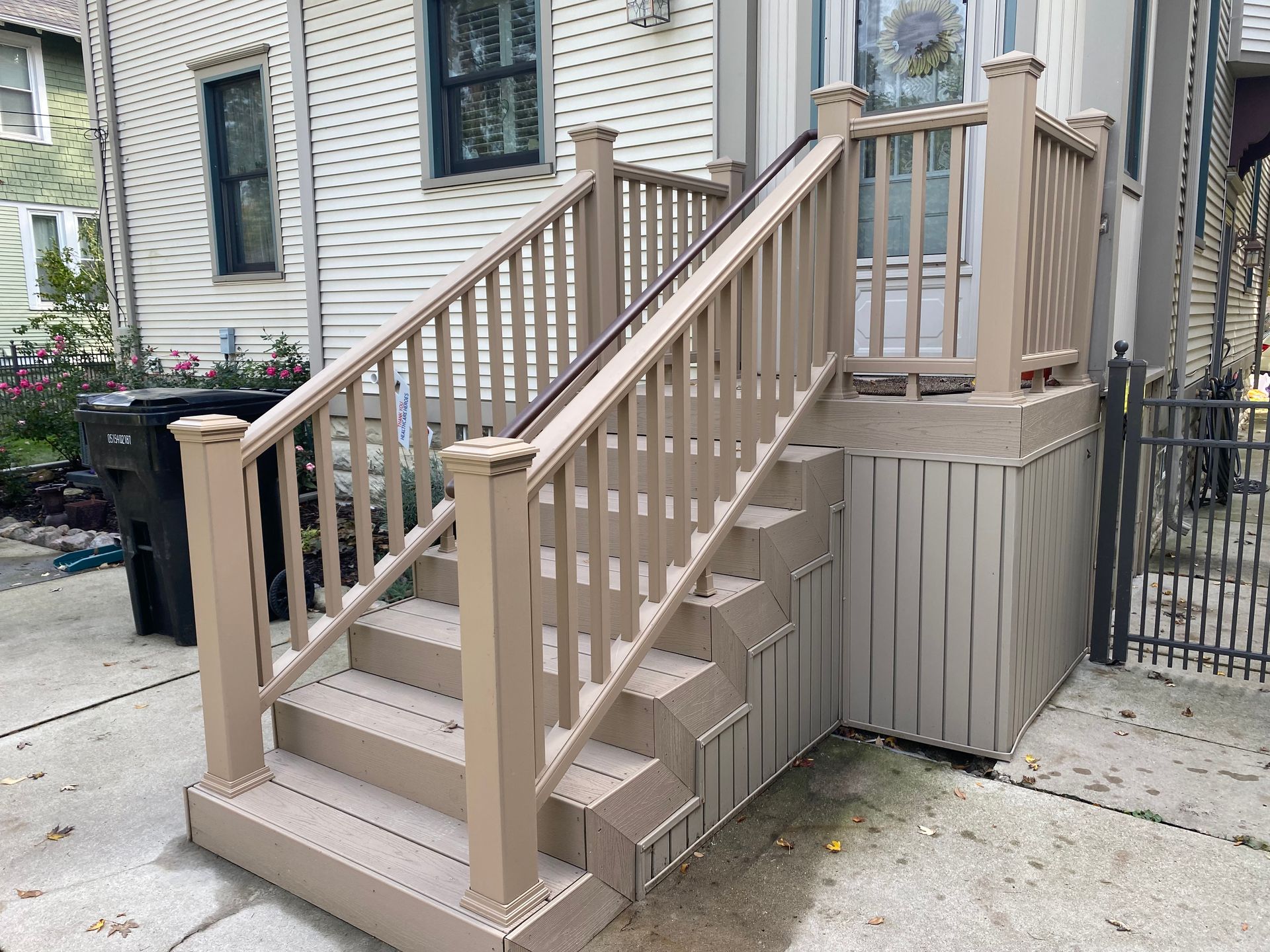 Beige deck and stairs leading to a doorway, with railings and a concrete base, next to a house.