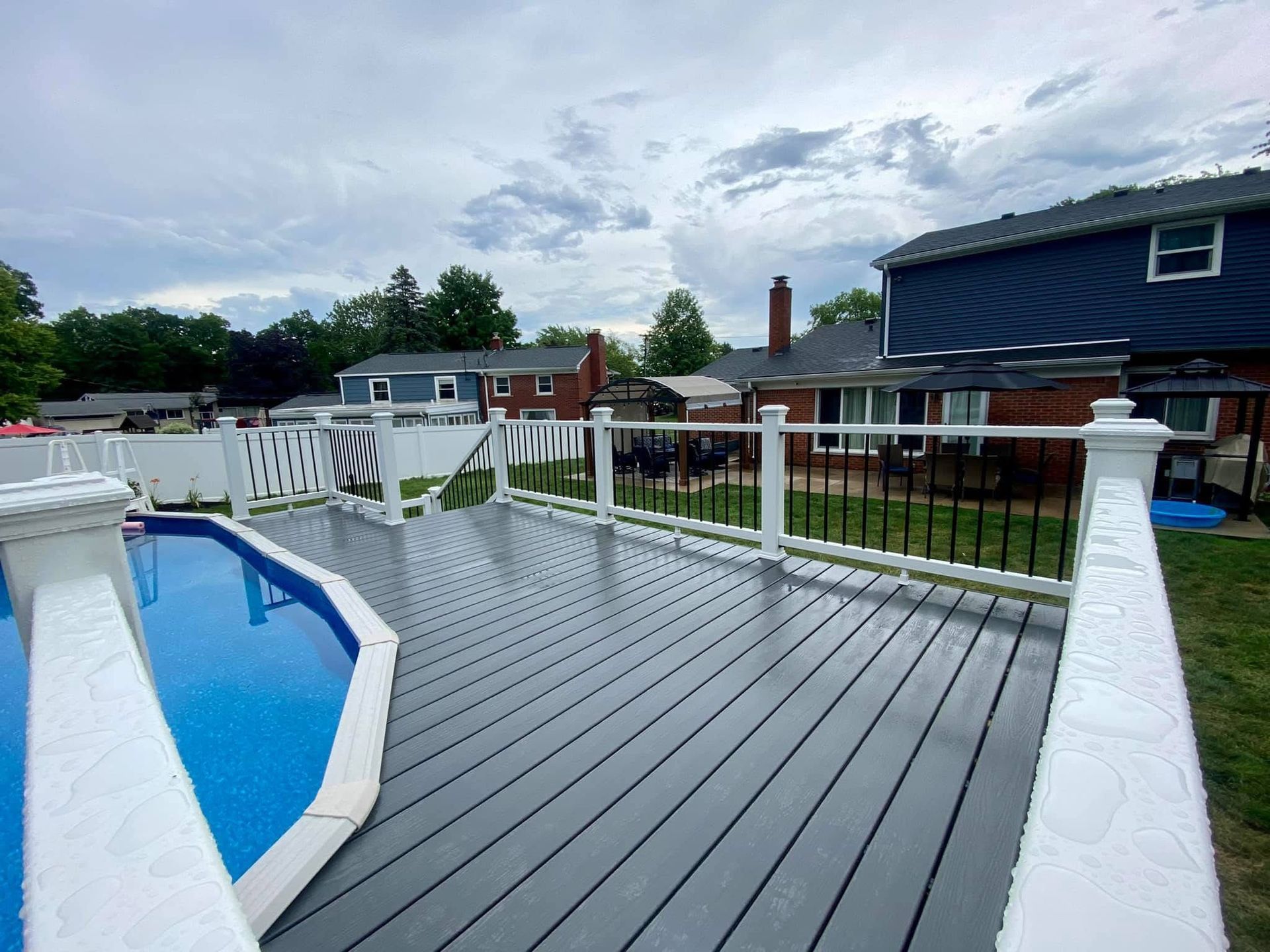 Grey composite deck with white railings, black metal balusters, and an above-ground pool under cloudy skies.