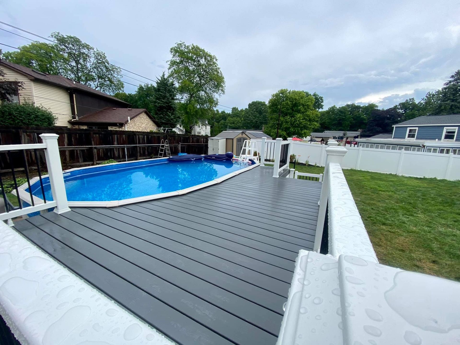 Above-ground pool with blue cover, gray deck, white railing, and grassy backyard on an overcast day.