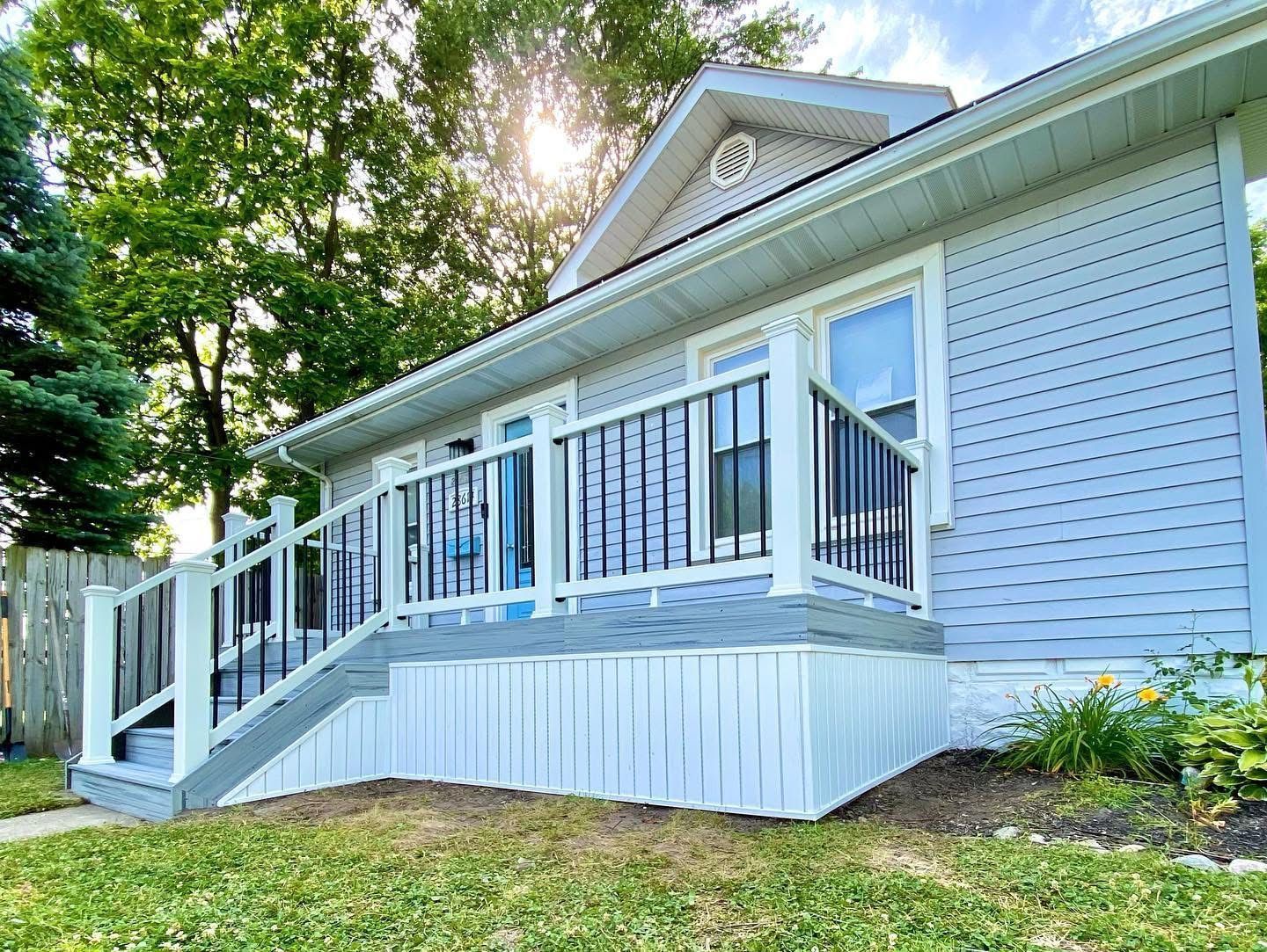 Light blue house with white trim, porch, and black railing; bright sun.