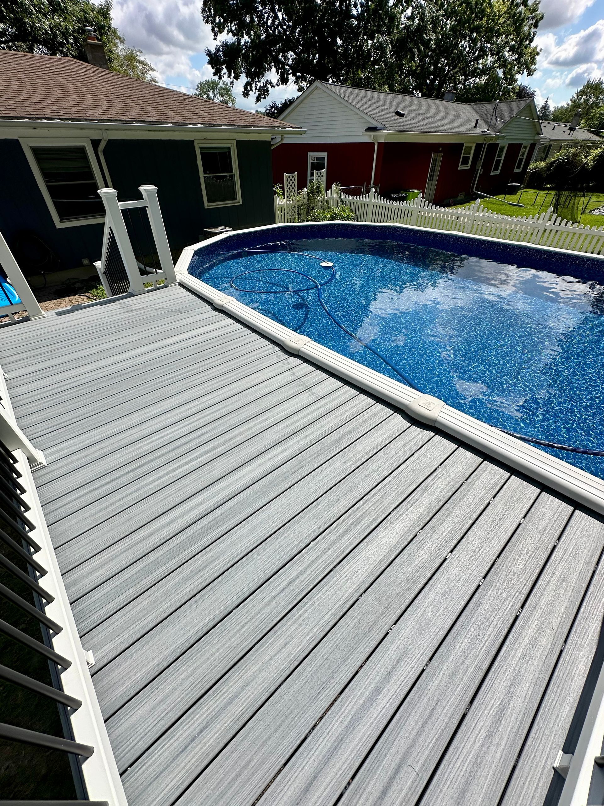 Gray deck next to an oval-shaped pool with blue water. Houses are in the background.