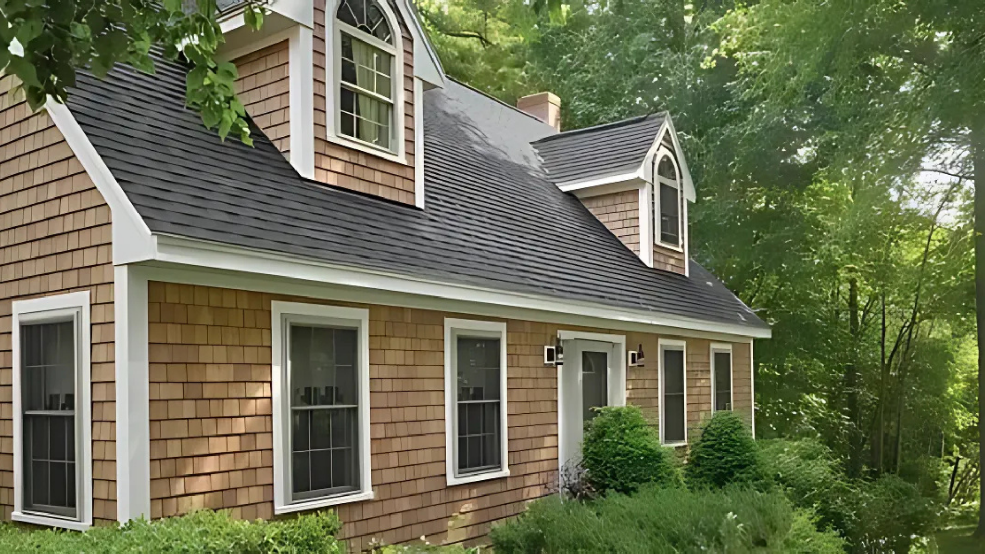 Tan shingled house with black roof and white trim, surrounded by green trees and bushes.