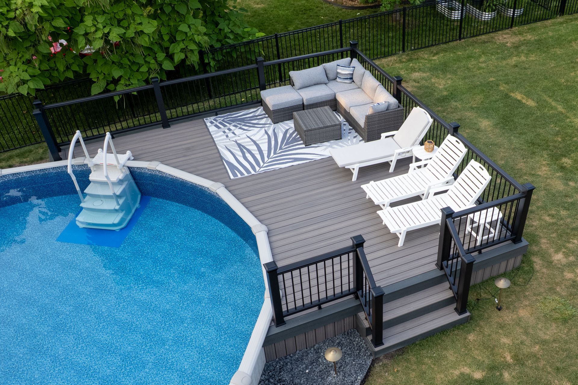Above-ground pool with deck, gray composite boards, black railing, outdoor furniture, and white lounge chairs.
