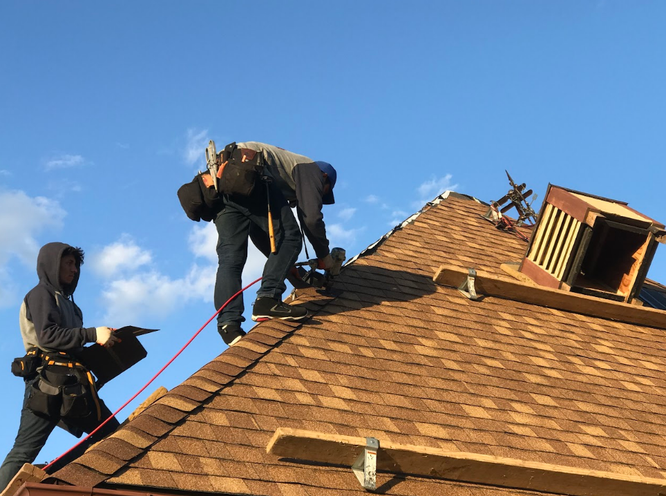 Two roofers working on a brown shingled roof under a blue sky, one attaching a new shingle.
