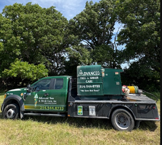 Green tow truck with “Advanced” signage parked on grass beside trees under a blue sky.