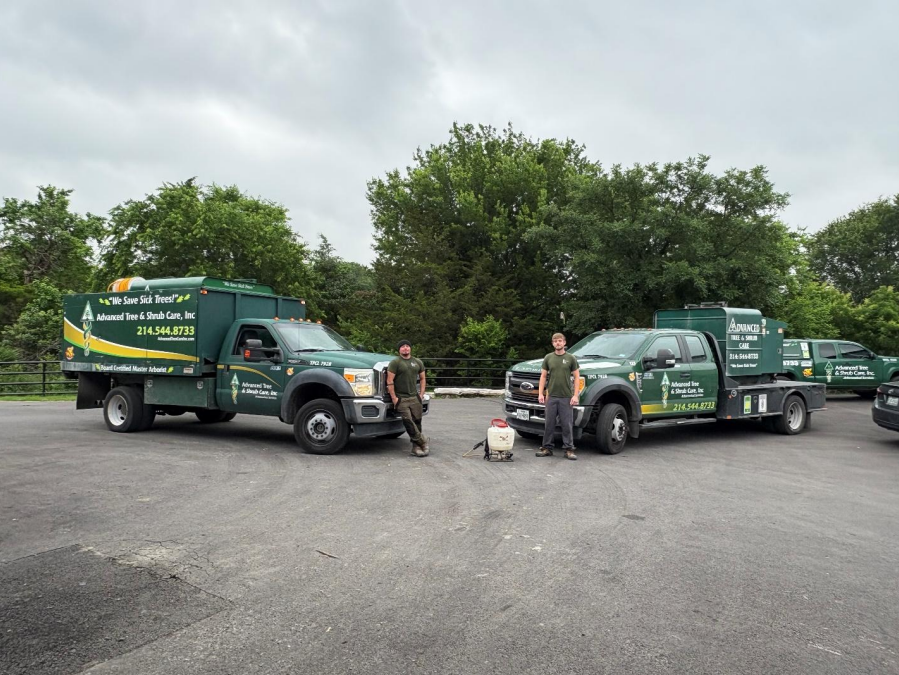 Two green utility trucks parked on a lot with workers standing nearby under cloudy skies