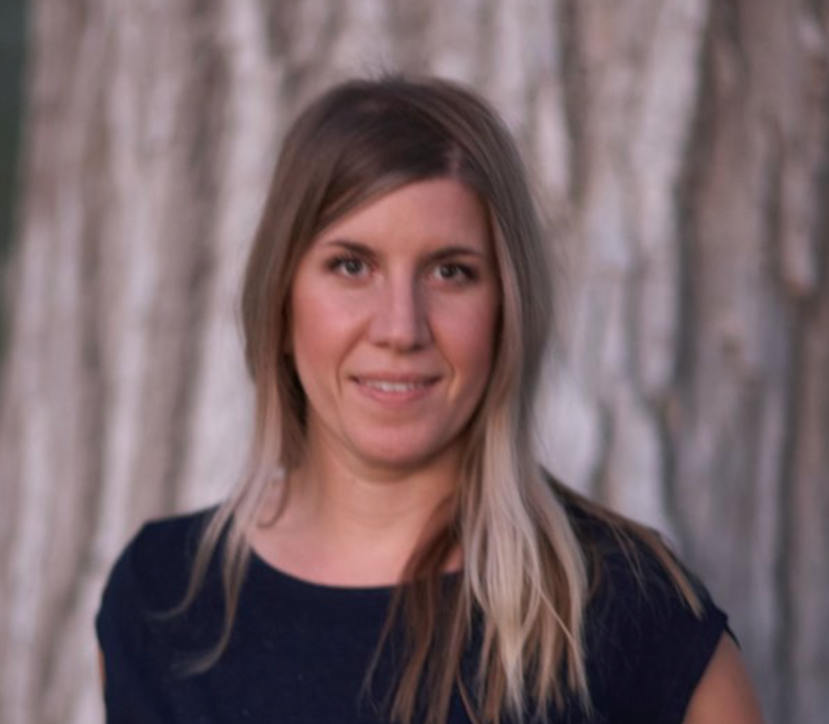 A woman in a blue shirt is smiling in front of a tree