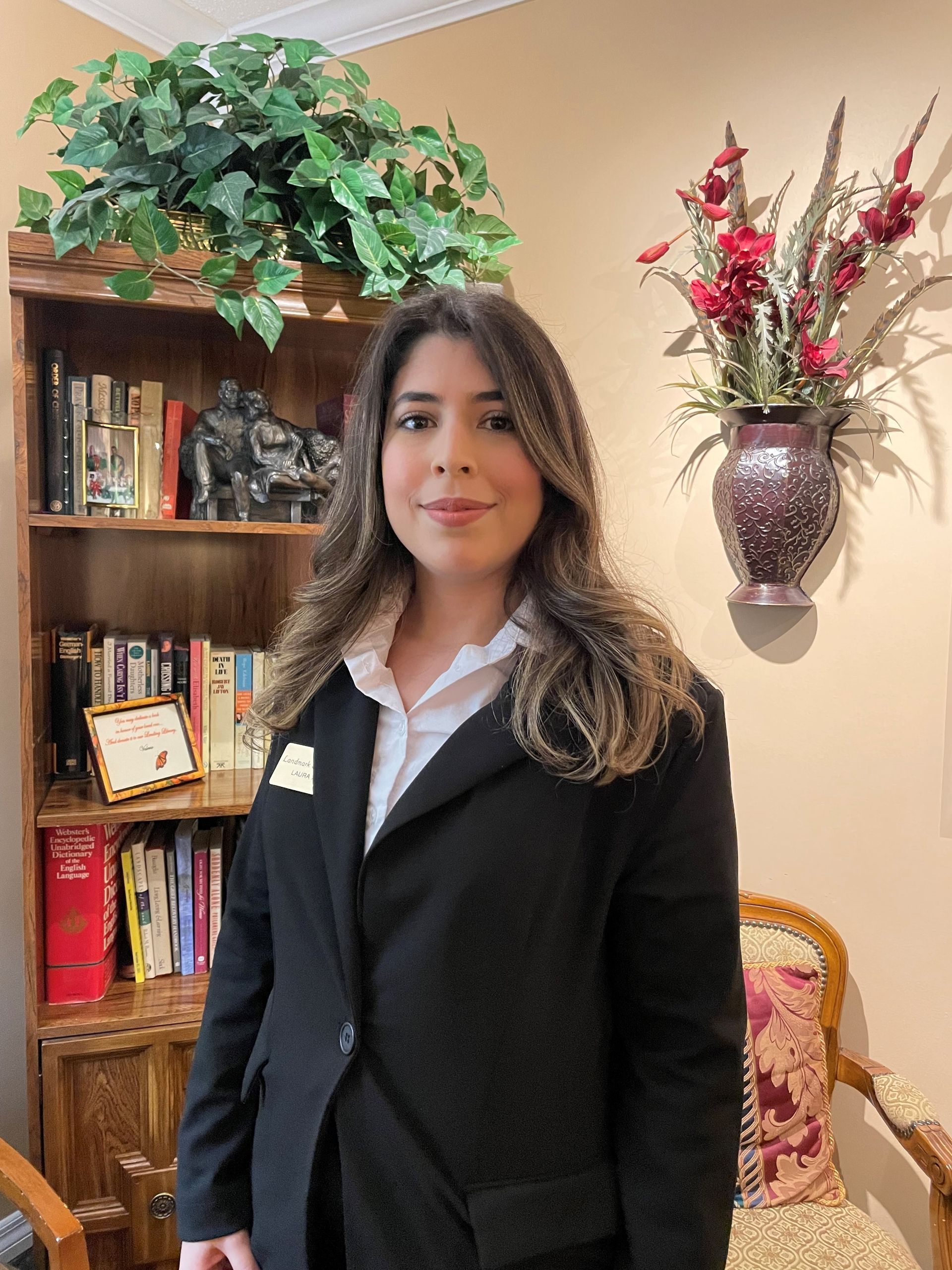 Woman in suit stands in front of a bookshelf and wall decor.