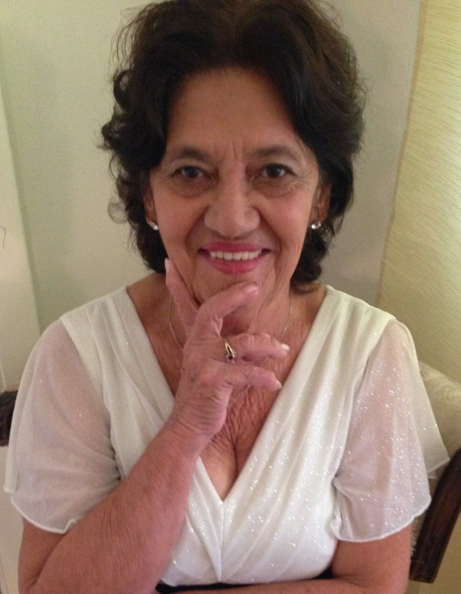 A woman in a white dress is smiling and holding her hand to her chin.