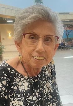 An elderly woman wearing glasses and a floral shirt is smiling for the camera.