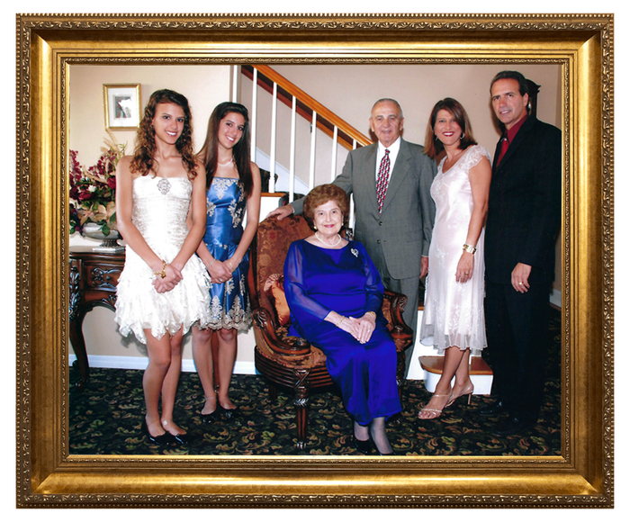 Family portrait in a gold frame: six people posing indoors; woman seated in chair, surrounded by others.