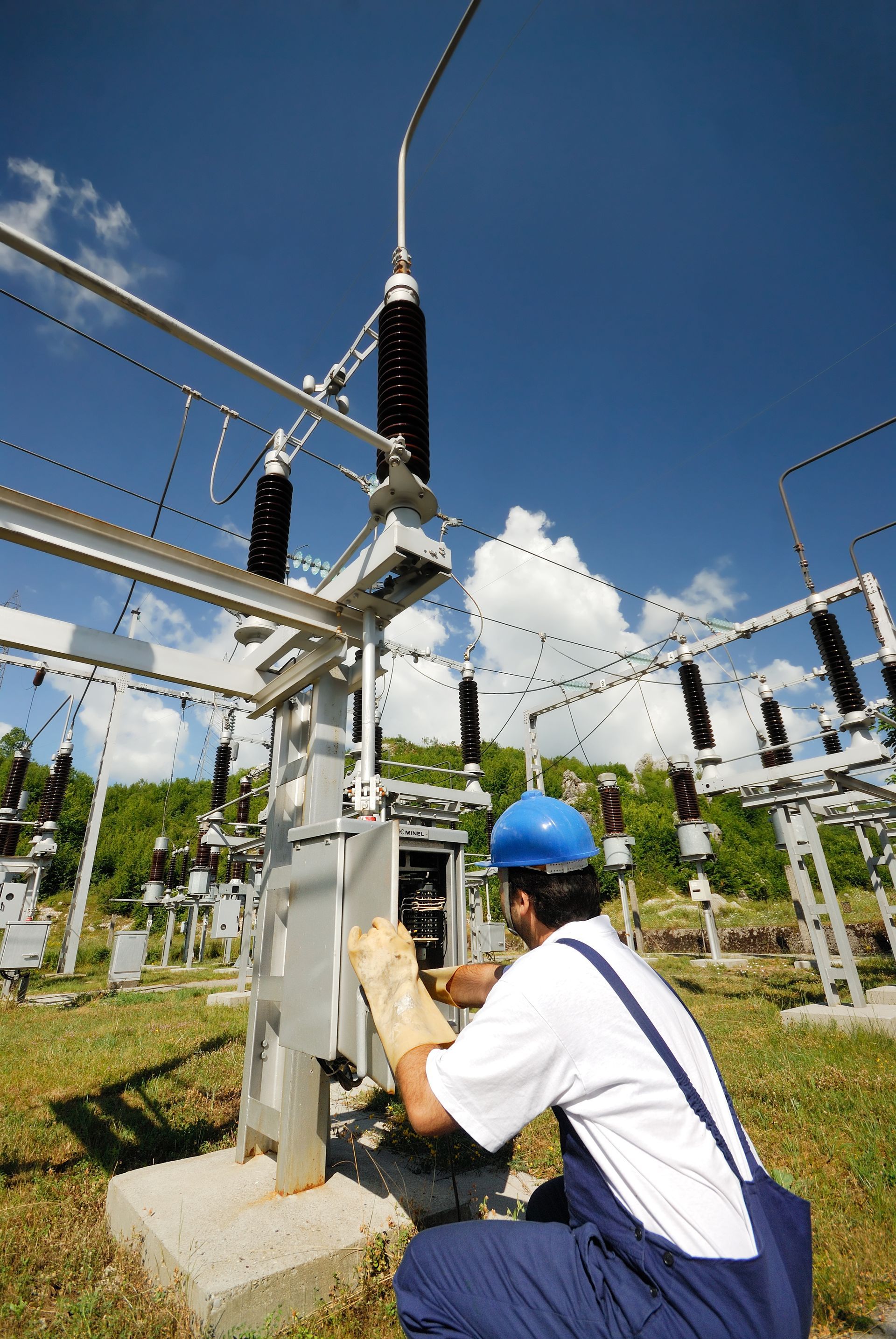 Technician in blue hard hat inspecting electrical substation equipment under a clear sky