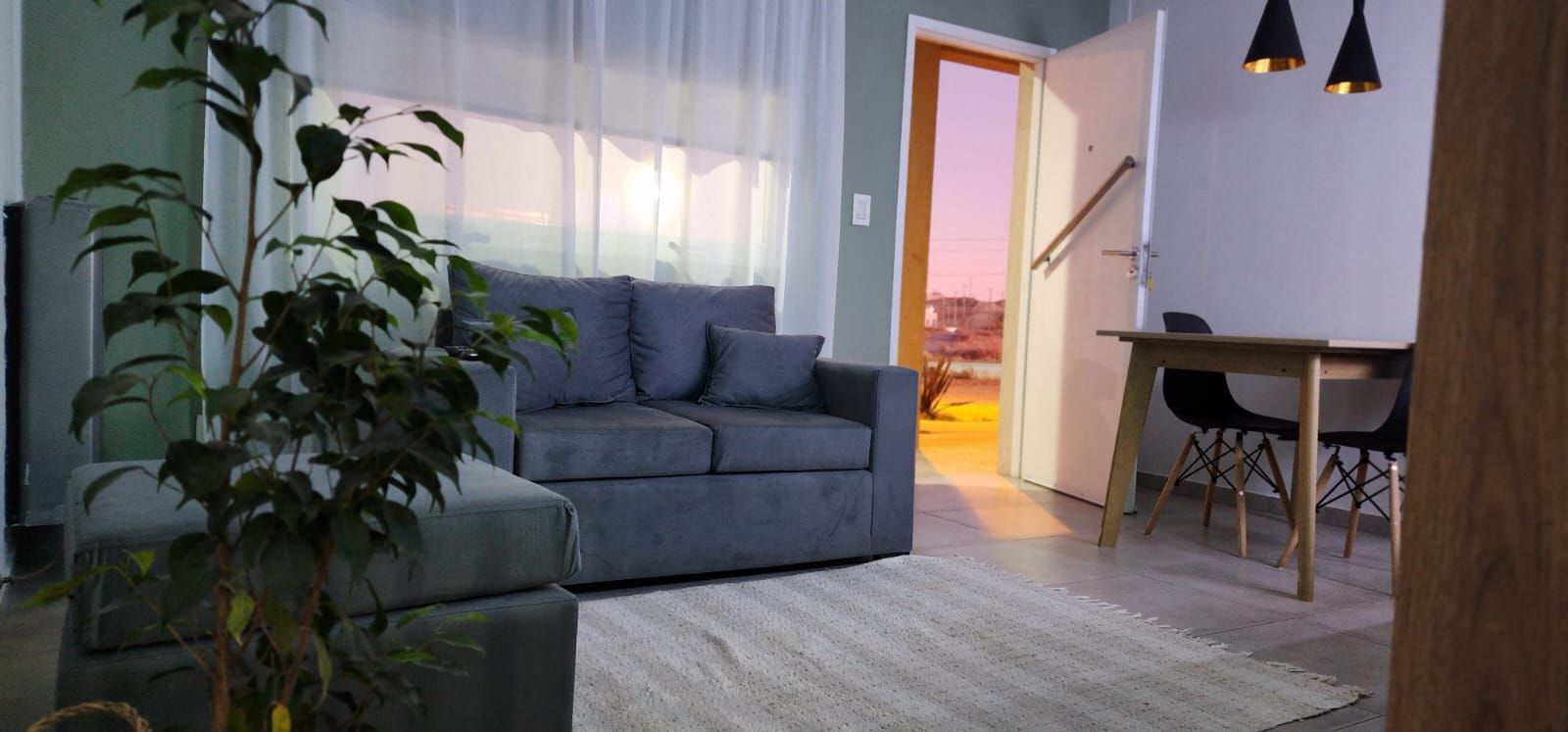 Sala de estar con sofá azul, mesa de comedor y una planta grande. La puerta da a una vista del atardecer.