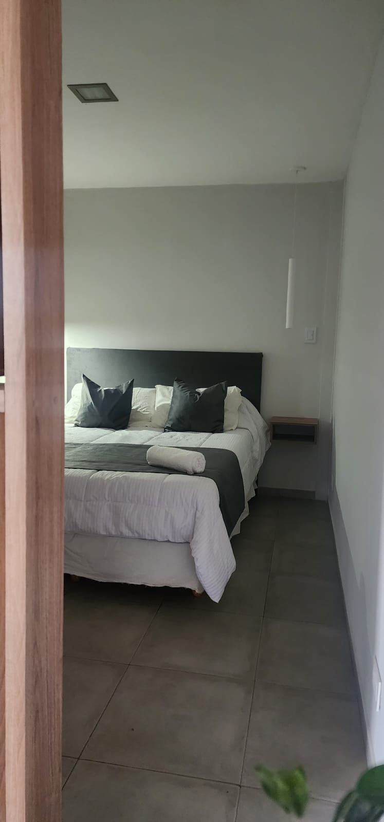 Interior de dormitorio con cama, almohadas y mesita de noche flotante. Paredes y suelo grises.