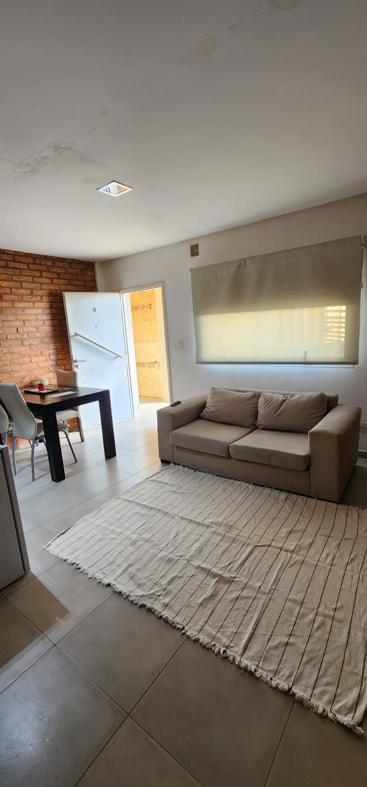 Una sala de estar con sofá, alfombra, mesa y pared de ladrillo. Puerta abierta al fondo.