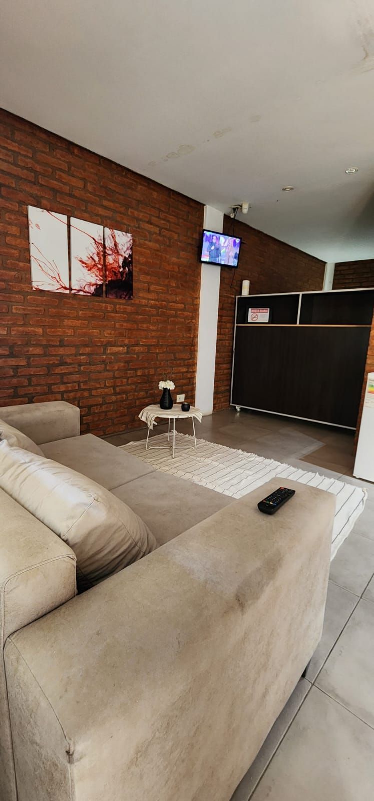 Una sala de estar con una pared de ladrillo, un sofá beige, un televisor y una pequeña mesa redonda.