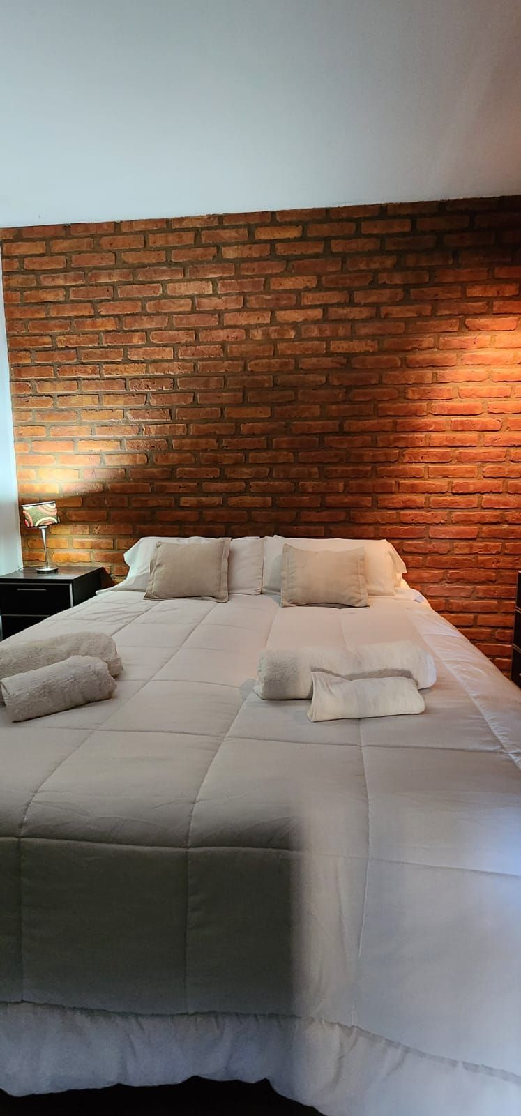Cama con ropa de cama blanca y toallas dobladas, contra una pared de ladrillos.