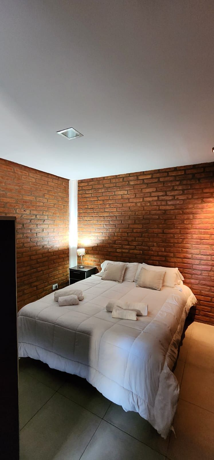 Dormitorio con paredes de ladrillo visto, cama blanca y toallas. Mesilla de noche negra con lámpara.