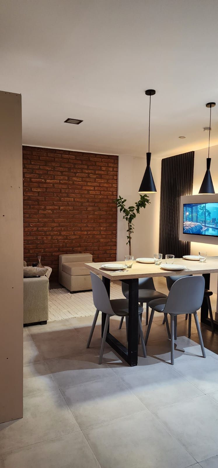 Comedor con mesa puesta para cuatro, pared de ladrillo, sillas grises, lámparas colgantes y TV.