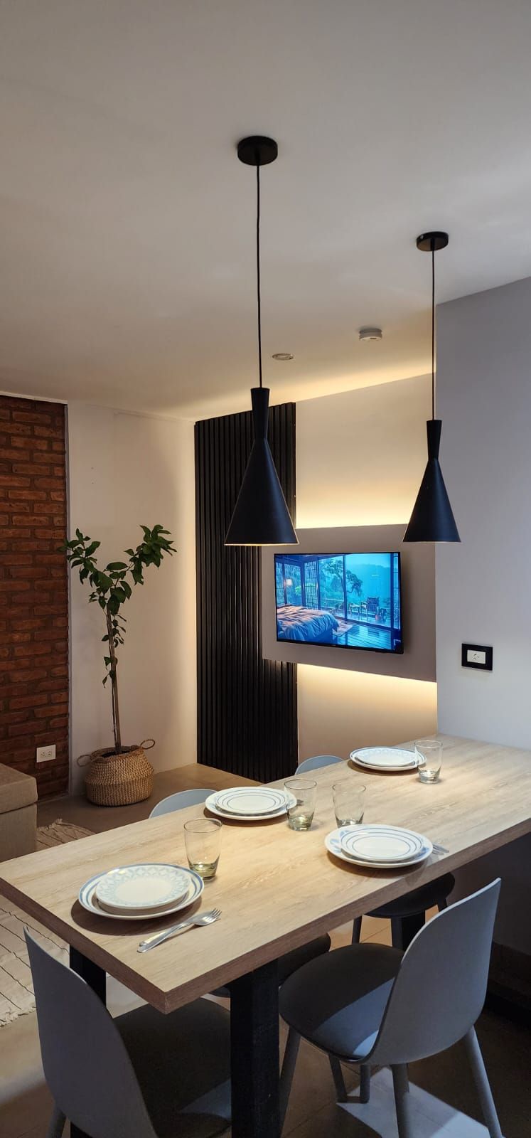 Comedor con mesa, sillas, lámparas colgantes, TV y pared de ladrillo.