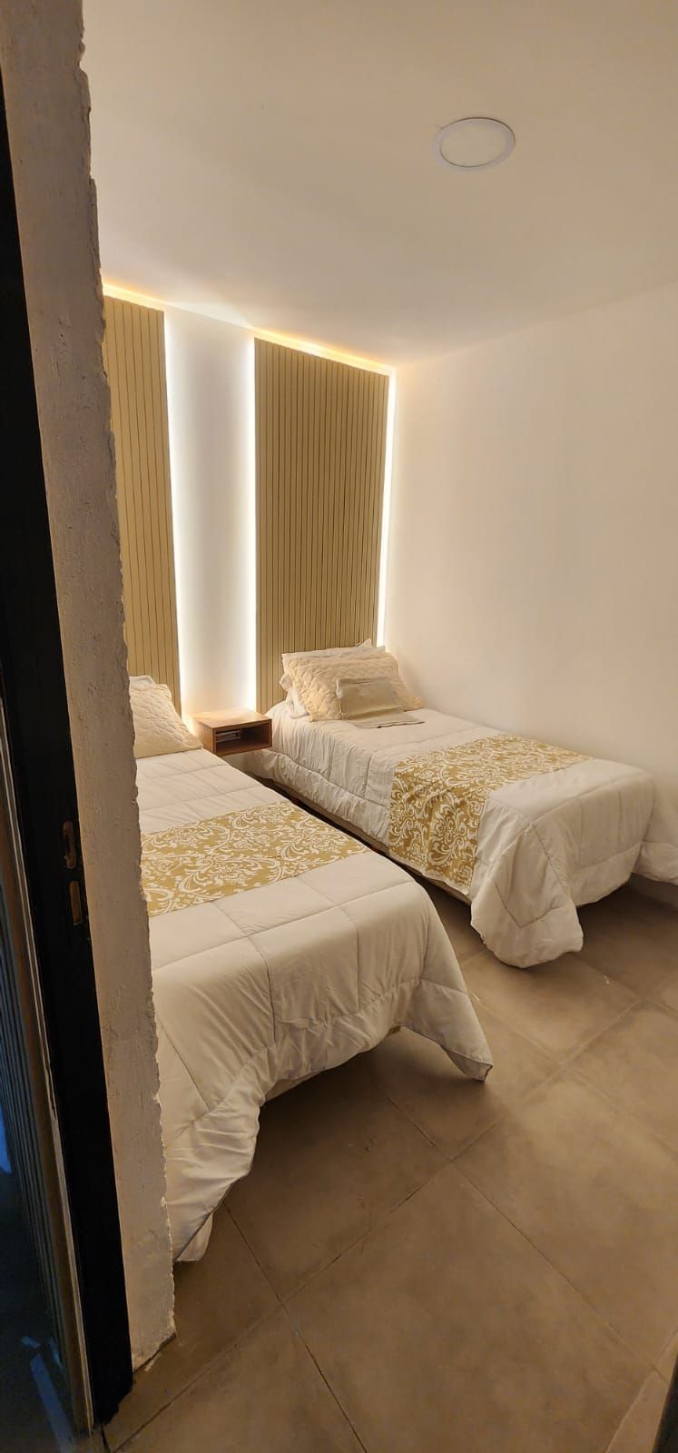 Dos camas individuales en una habitación moderna con colores neutros, pared decorativa detrás de ellas e iluminación cálida.