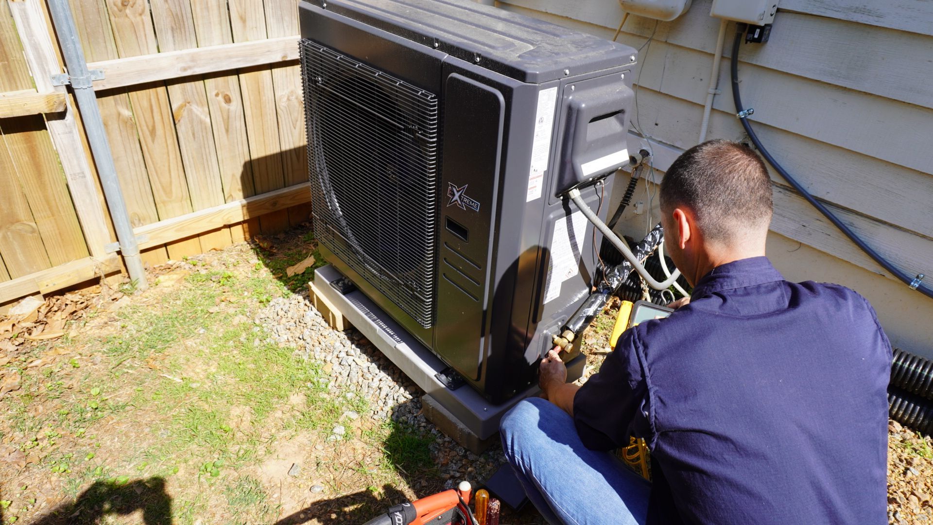 Technician working beside an outdoor HVAC unit near a brick wall, inspecting it with tools nearby