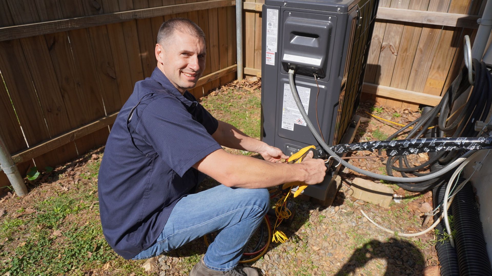 Technician crouches by an outdoor electrical panel, working on cables beside a wooden fence.